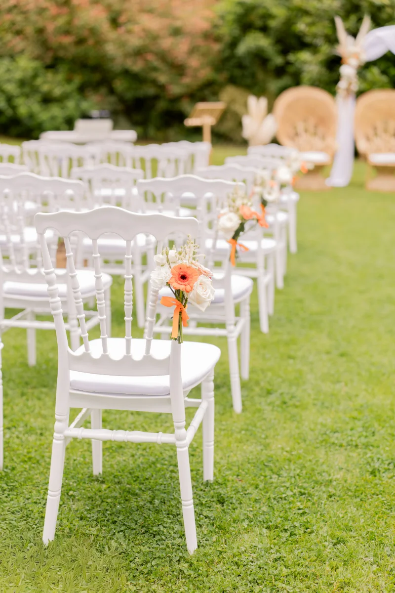 Allée de cérémonie sur la pelouse : chaises Napoléon blanches ornées de bouquets orange, fauteuils en rotin au fond