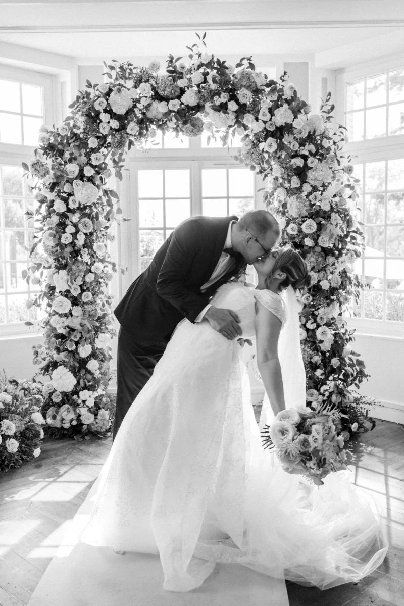 Baiser renversé des mariés sous une arche de fleurs blanches, dans une salle lumineuse aux grandes fenêtres, en noir et blanc