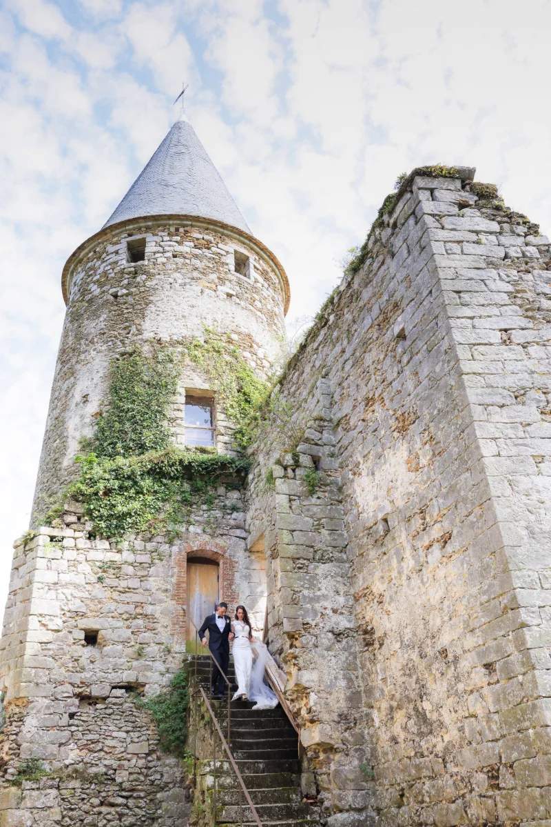 Les mariés descendent l'escalier d'une tour médiévale en pierre couverte de lierre, sous un ciel nuageux