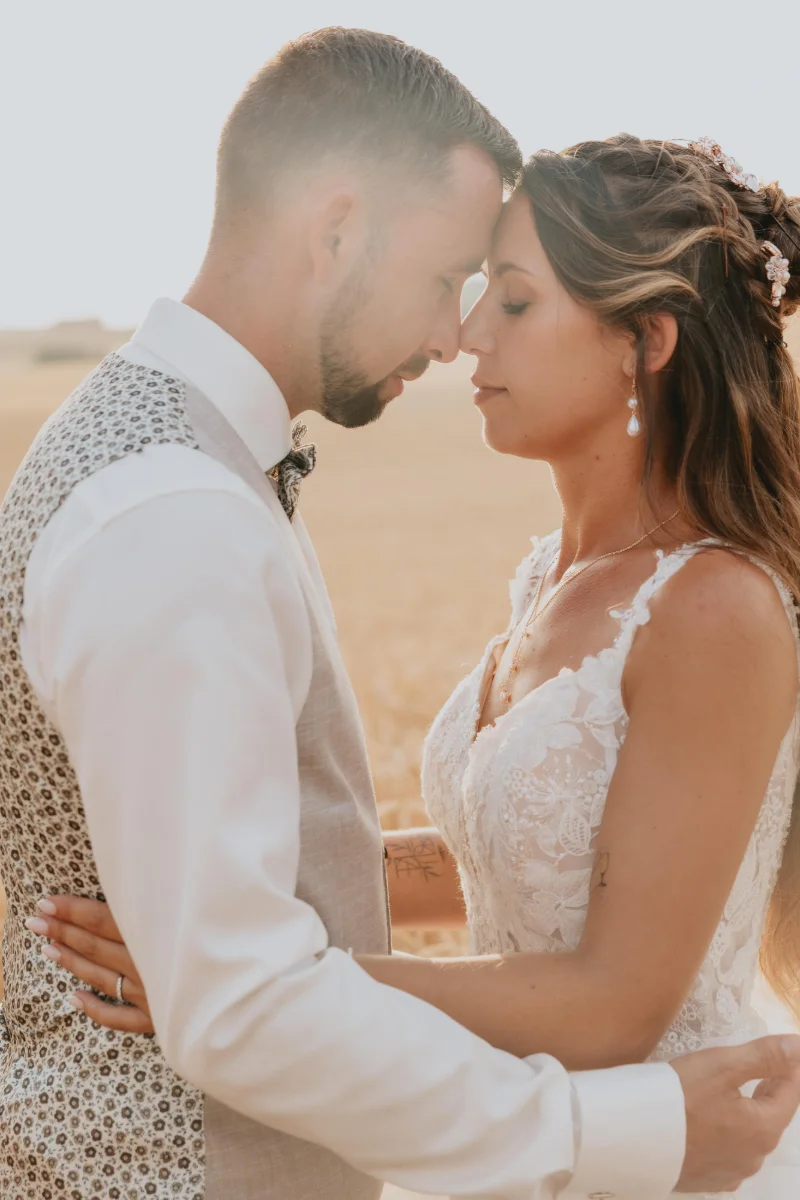 Les mariés front contre front dans la lumière dorée, gilet à motifs fleuris et robe de dentelle