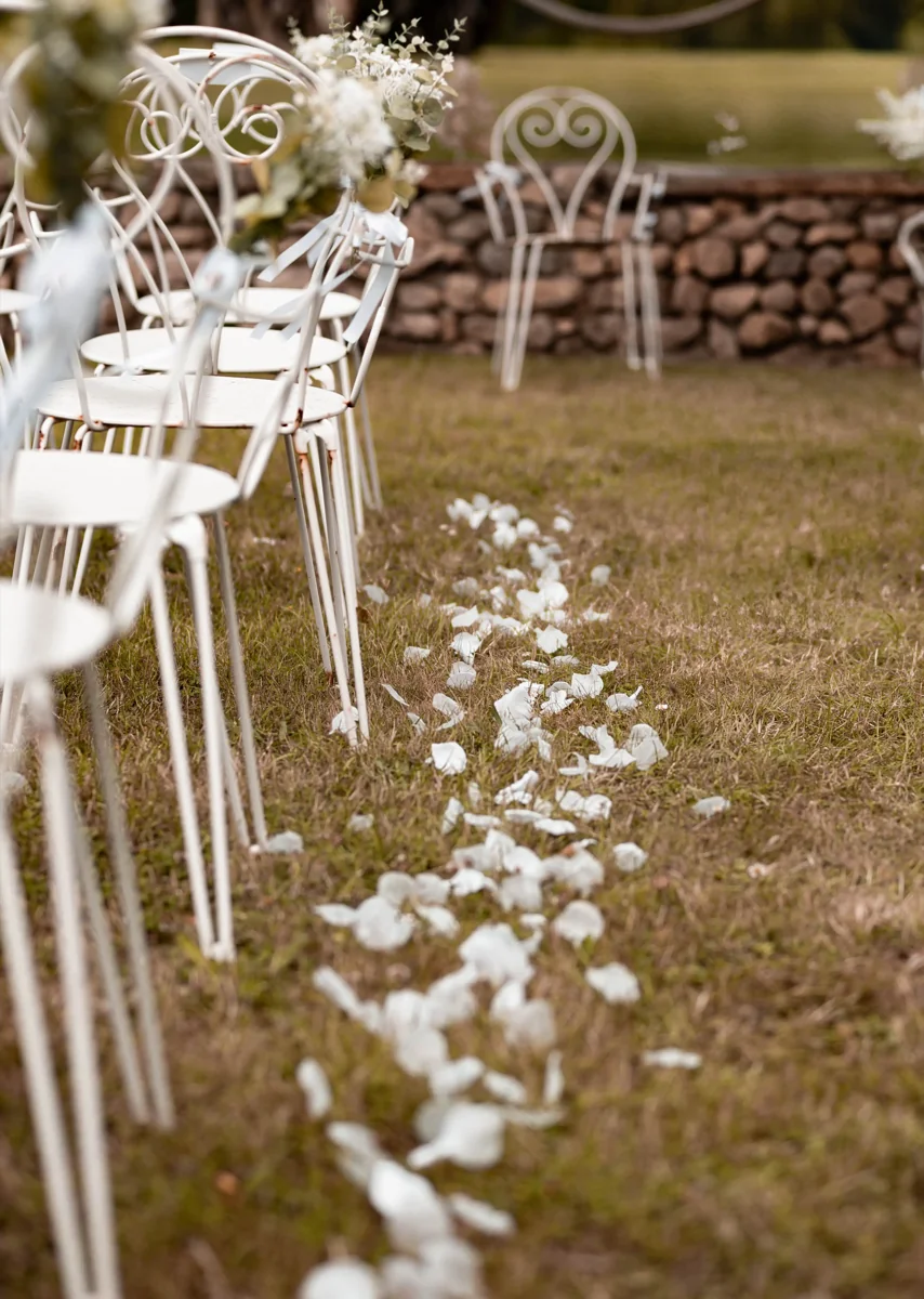 Allée de cérémonie parsemée de pétales blancs, chaises en fer forgé blanc ornées de petits bouquets sur l'herbe
