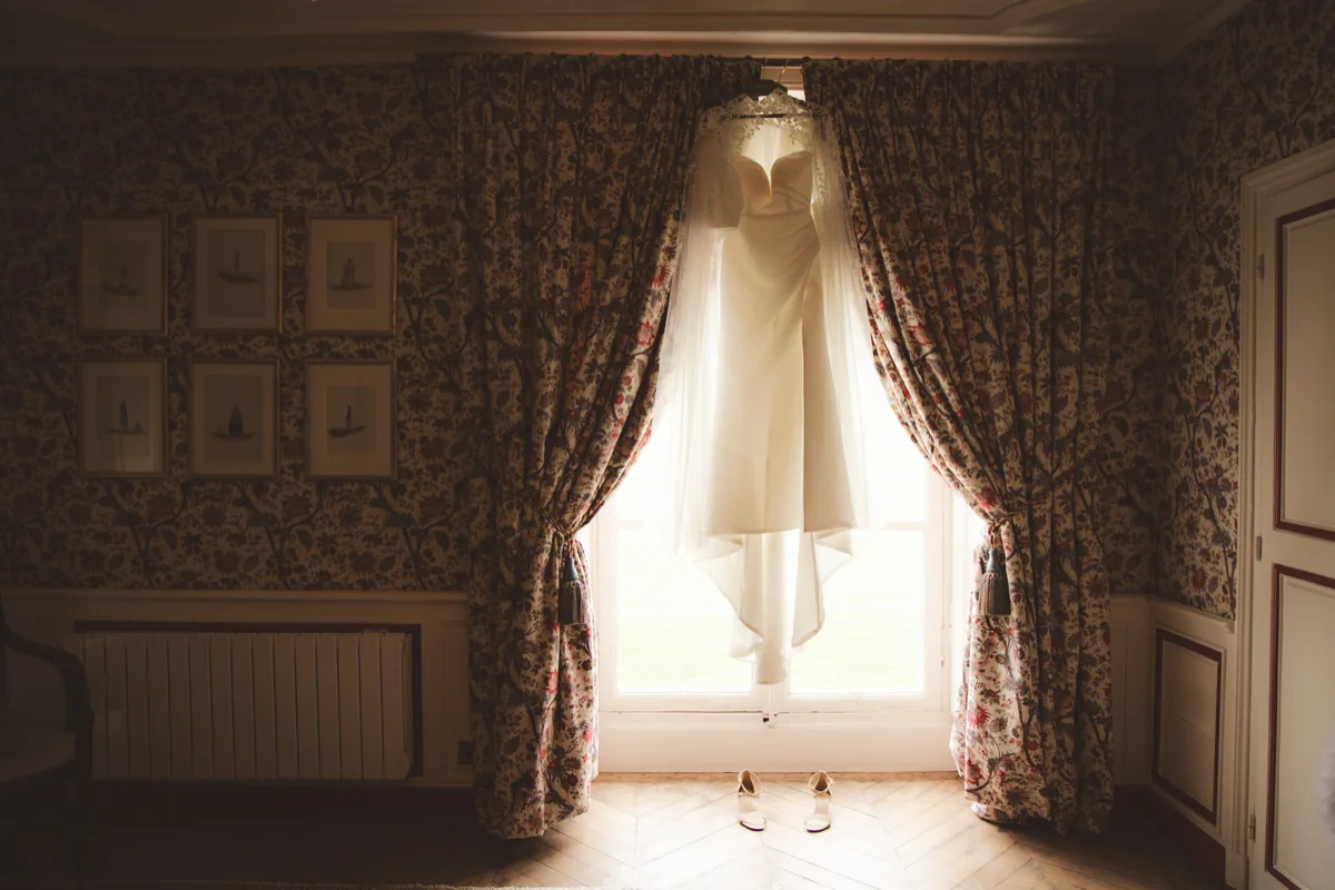 Robe de mariée suspendue devant une fenêtre lumineuse, rideaux fleuris, chaussures posées sur le parquet en chevrons