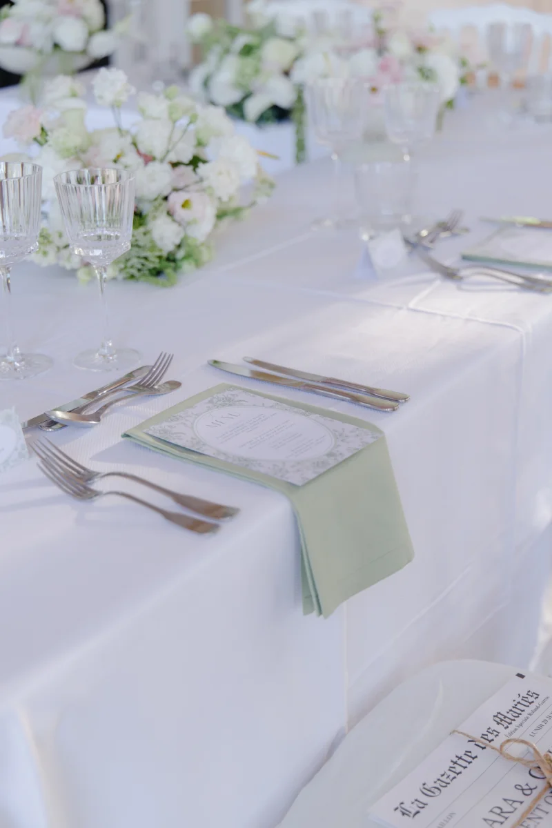 Détail d'une table de mariage : nappe blanche, serviette vert sauge, menu et verres en cristal ciselé