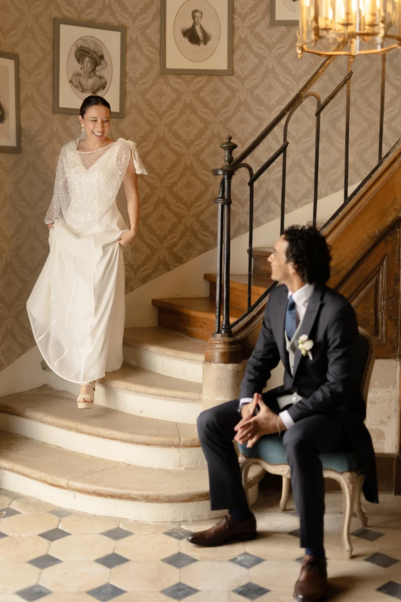 La mariée descend l'escalier en riant vers le marié assis qui l'attend, dans un hall au sol en damier