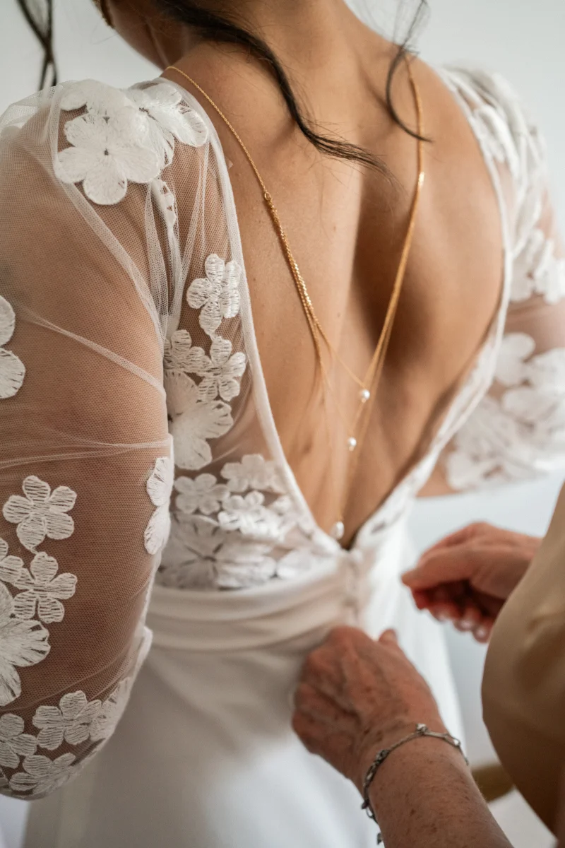 Préparatifs : une main boutonne le dos de la robe en dentelle fleurie, un collier en chaîne dorée sur le dos de la mariée